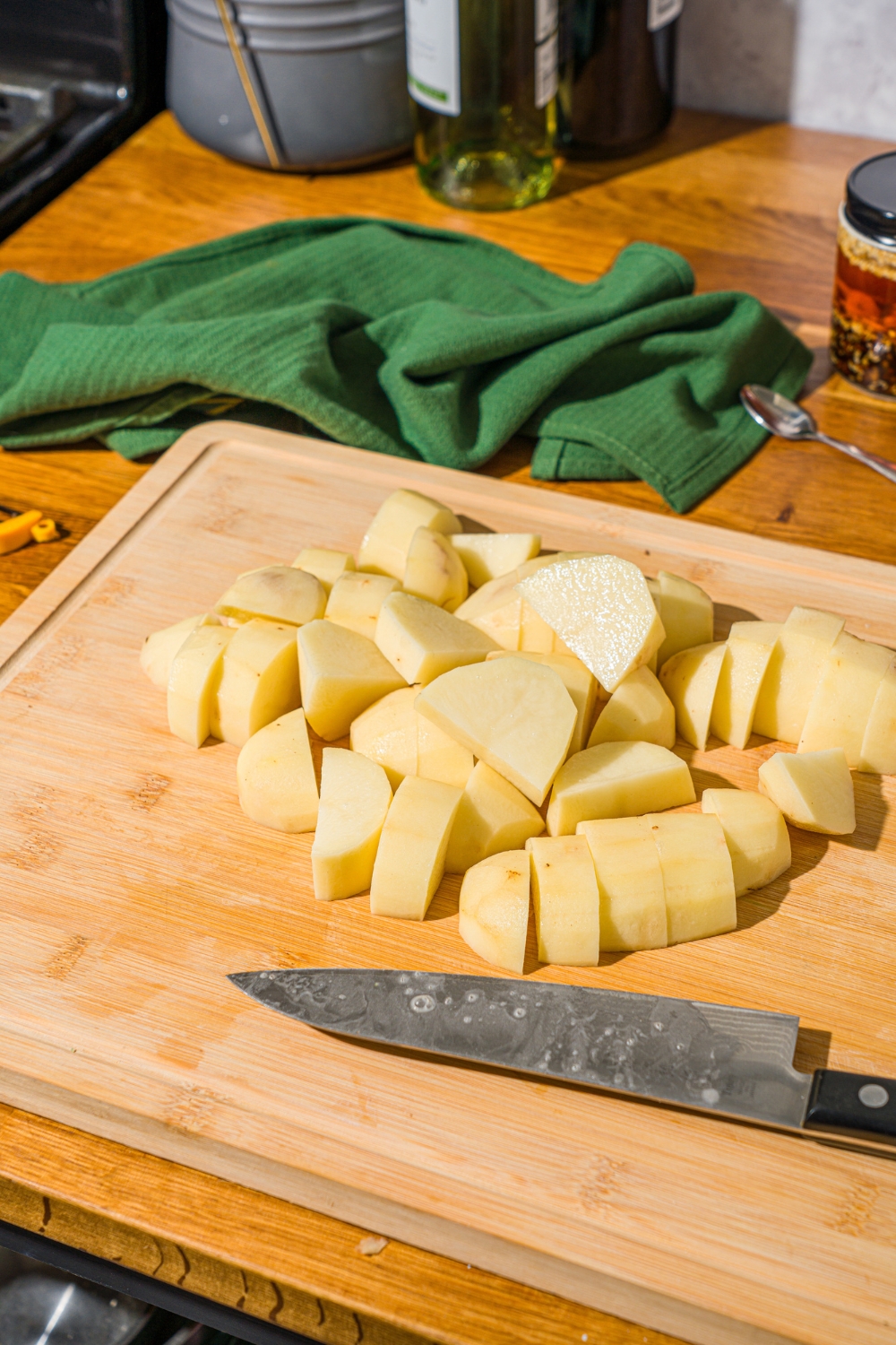 A cutting board with cubed potatoes and a large knife. The board is on a wooden counter with a green cloth napkin.