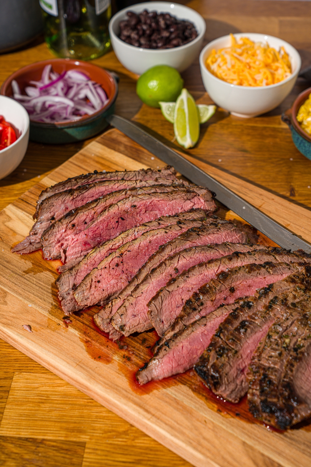 A cutting board with grilled marinated steak sliced thin. The board is on a wooden counter with several bowls of toppings.