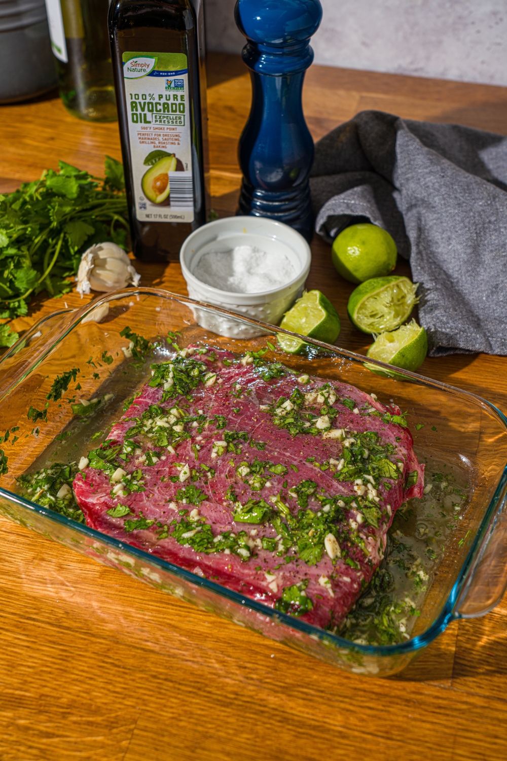 A glass baking dish with steak marinating in a cilantro lime marinade. The dish is on a wooden counter with lime, olive oil, cilantro, and a blue cloth napkin.