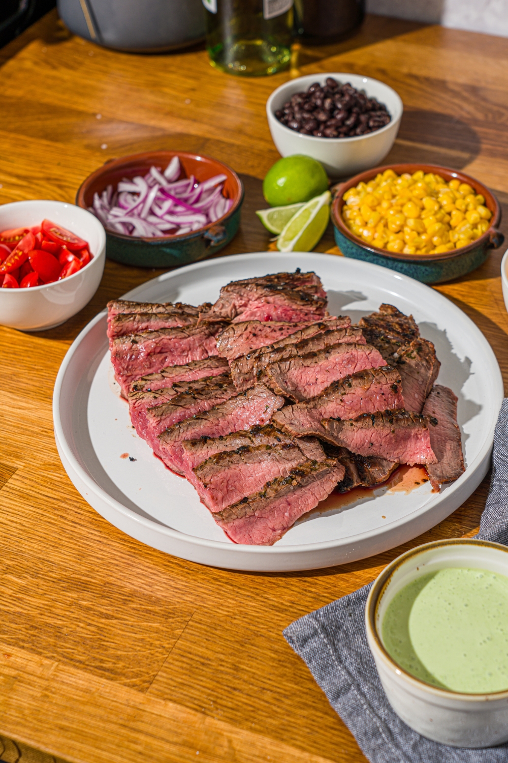 A white plate with marinated grilled steak sliced thin. The plate is on a wooden counter with several bowls of toppings.