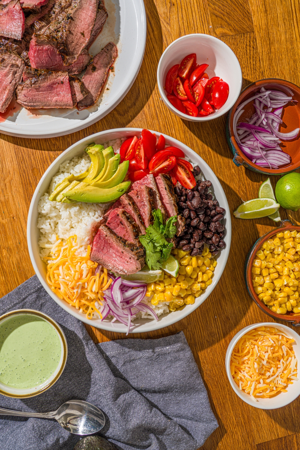 A large white bowl with a steak bowl served with rice, grilled marinated steak, corn, avocado, beans, tomato, red onion, and shredded cheese. The bowl is on a wooden counter with several bowls of toppings.