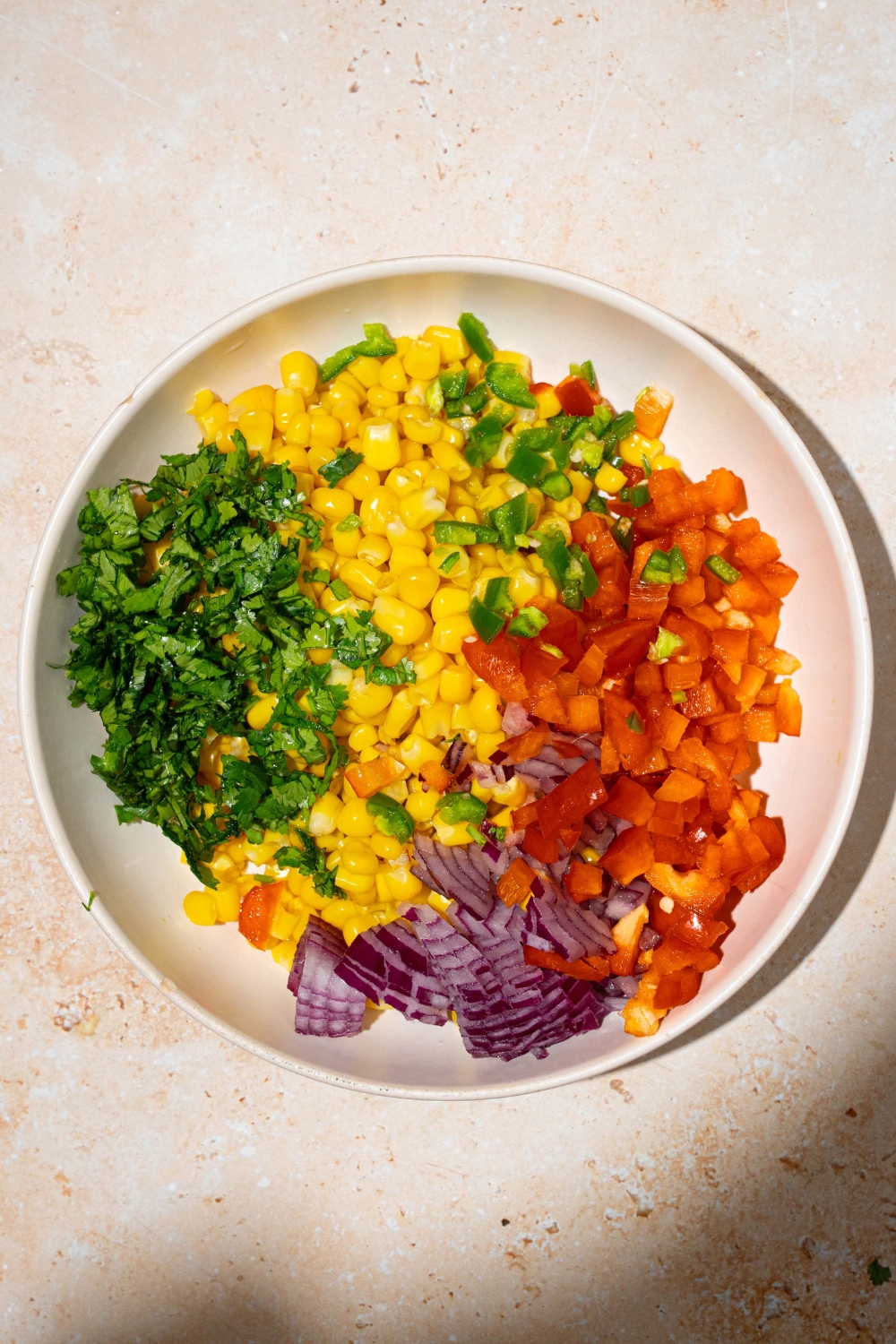 A white bowl with corn, chopped red pepper, chopped red onion, chopped cilantro, and chopped jalapeño. The bowl is on a tan counter
