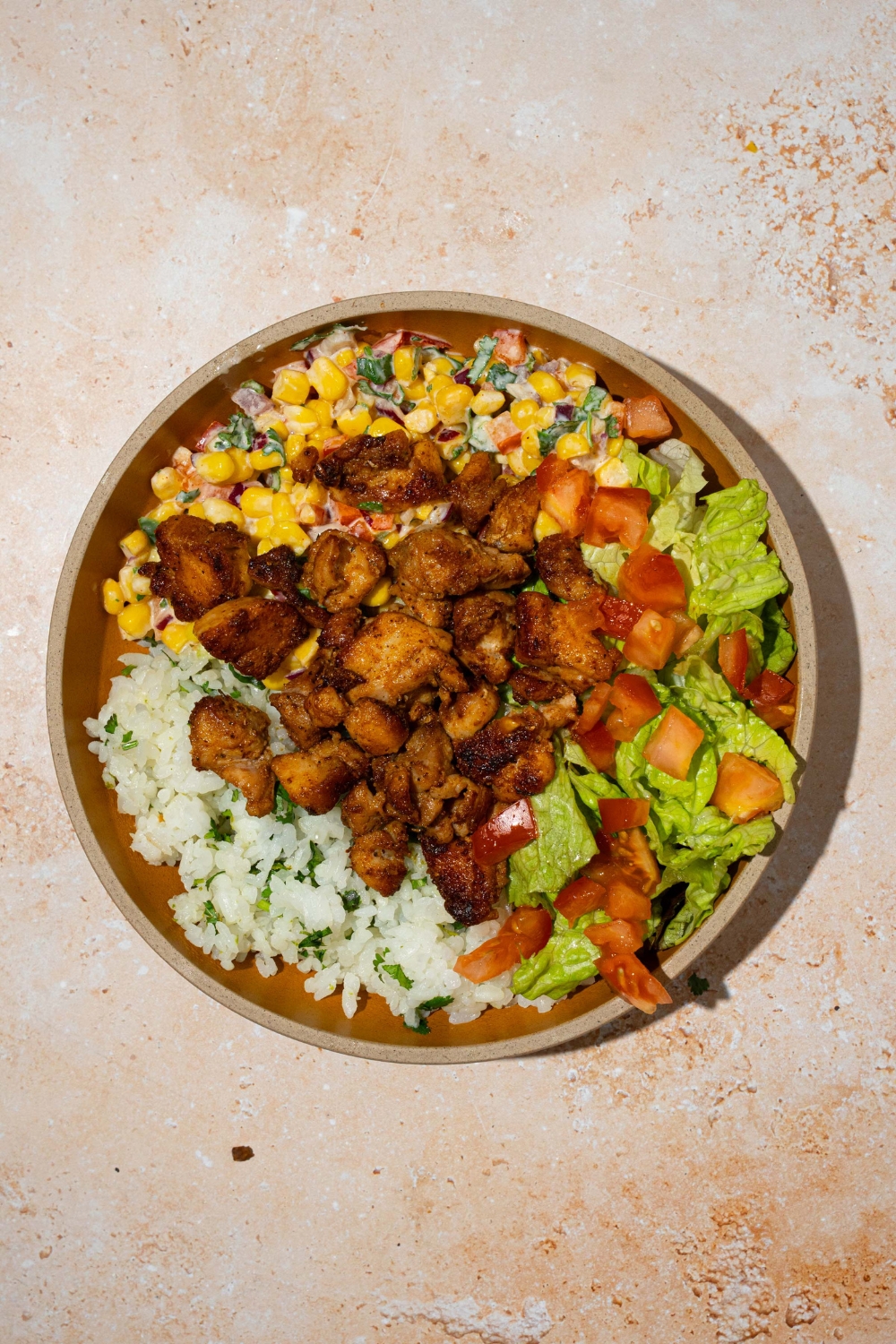 A brown plate with a street corn chicken rice bowl with cilantro lime rice, seasoned chicken, street corn mixture, lettuce, and tomatoes. The plate is on a tan counter.
