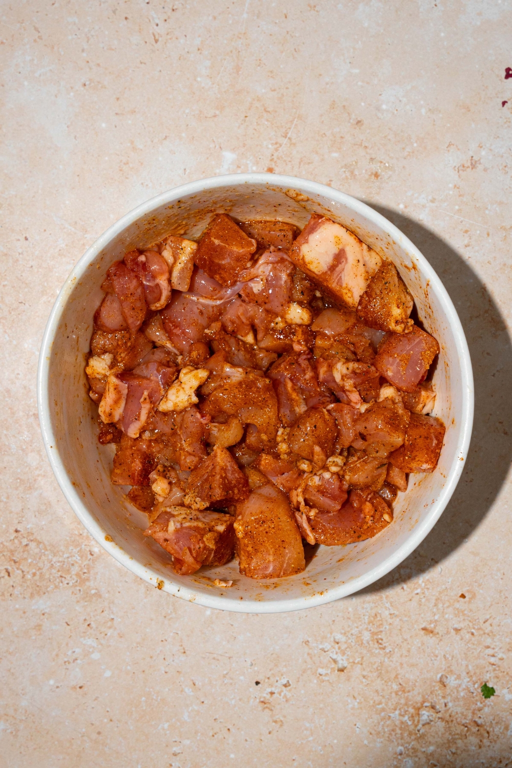 A white bowl with cubed chicken tossed in oil and seasonings. The bowl is on a tan counter.