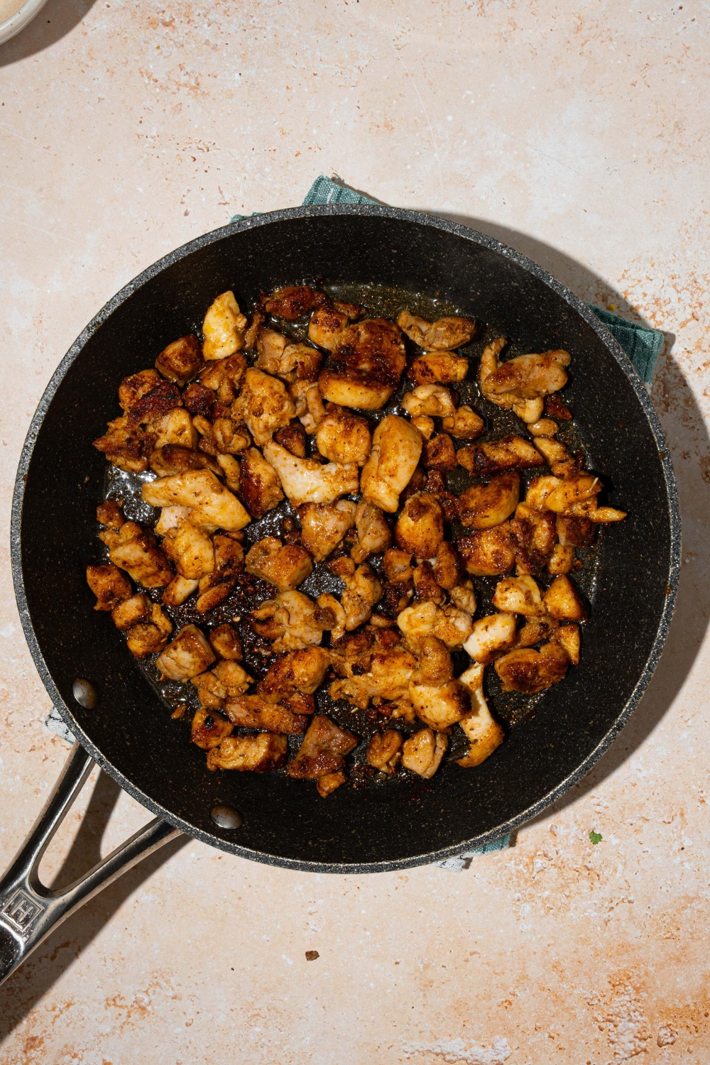 A skillet with seasoned chicken cooked in oil. The skillet is on a tan counter.