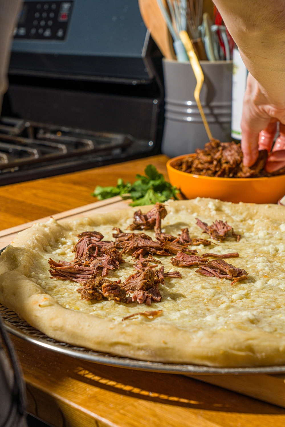 A half baked pizza on a pizza tray being topped with birria meat. The tray is on a wooden counter.