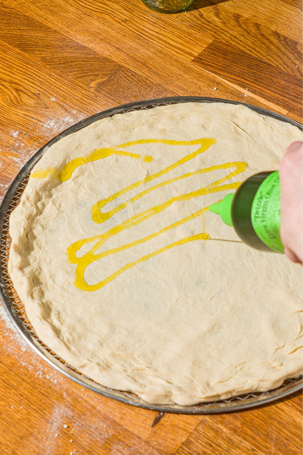 A pizza tray with rolled pizza dough being drizzled with oil. The tray is on a wooden counter.