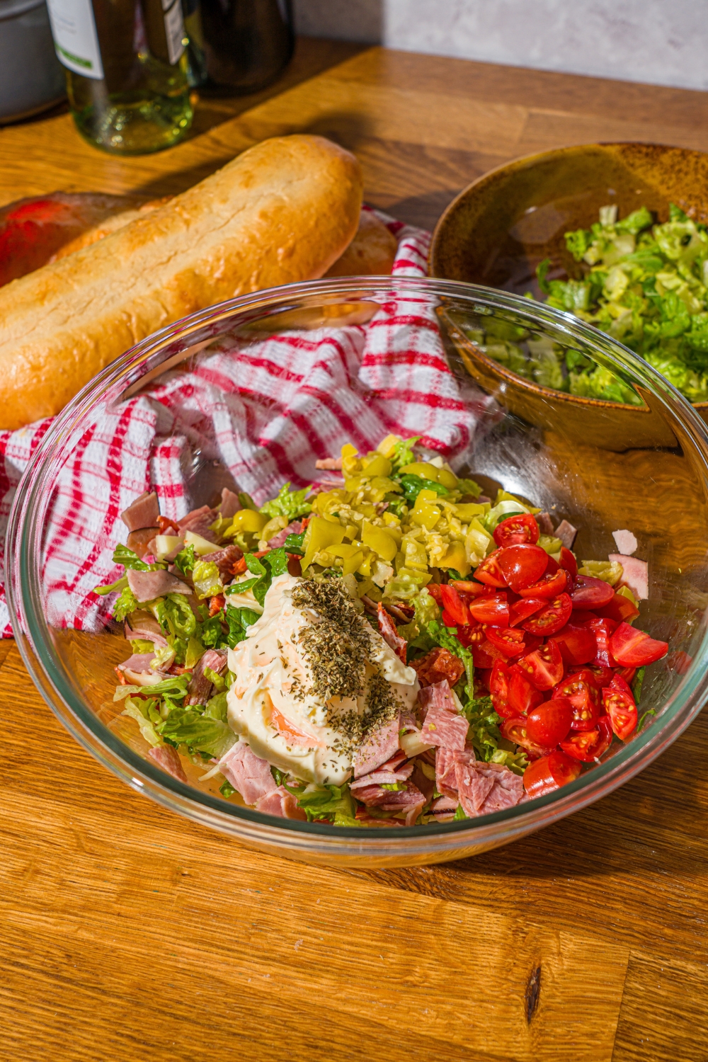 A glass bowl with chopped meats, tomatoes, lettuce, pepperocini, mayo, and seasonings. The bowl is on a wooden counter with a red striped napkin.