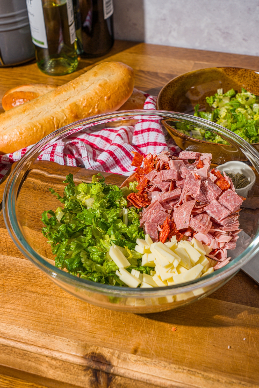 A glass bowl with chopped lettuce, meats, and provolone cheese. The bowl is on a wooden counter with a red striped napkin.