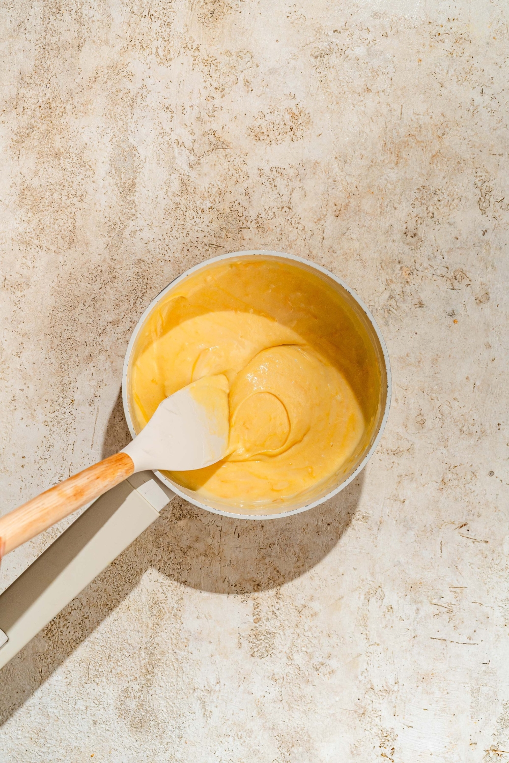 A spatula mixing a small bowl of cheese sauce. The bowl is on a tan counter.