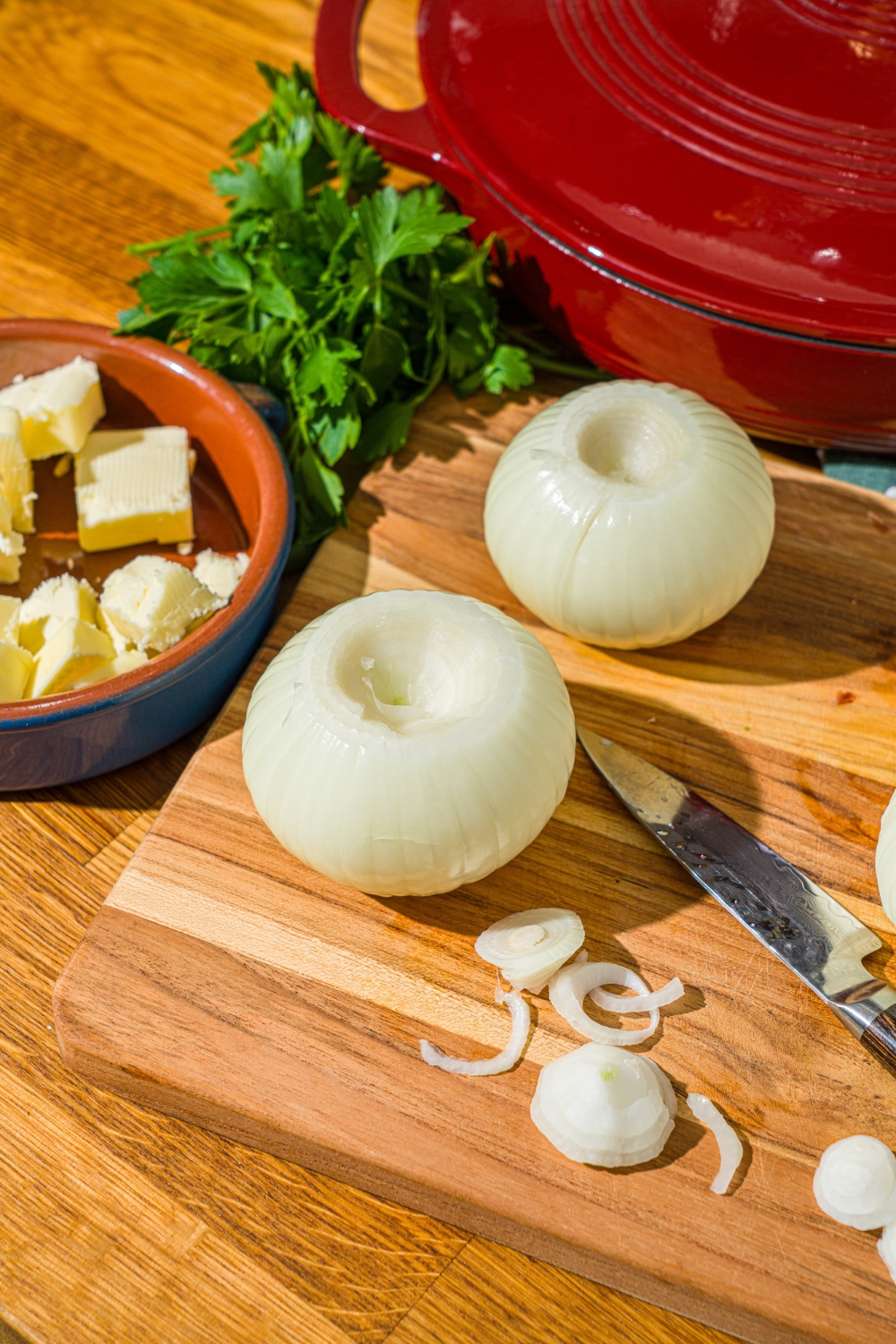 A cutting board with a knife and onions with the tops sliced off. The board is on a wooden counter with a dutch oven and bowl of butter.