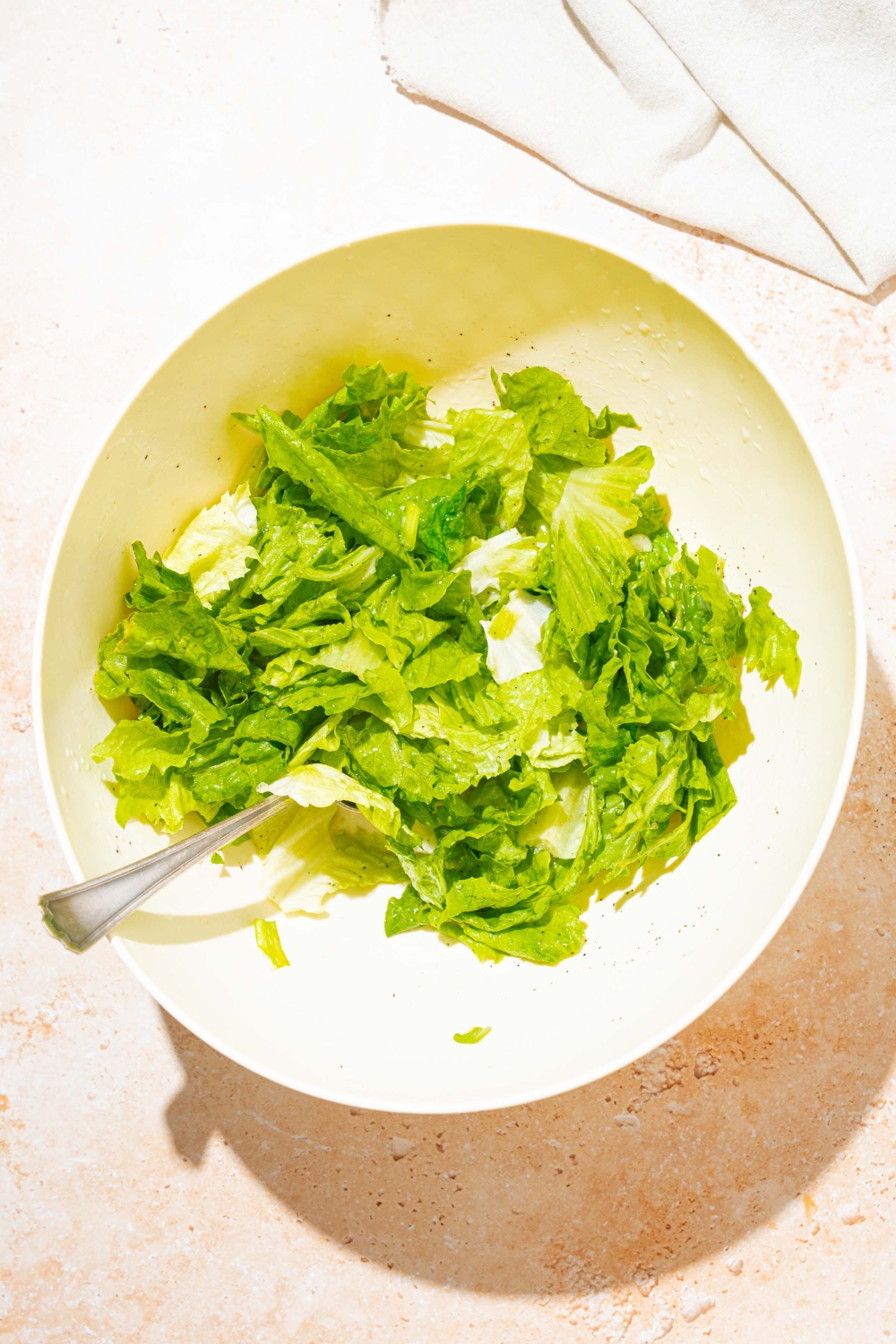 A large white bowl with chopped lettuce and a fork tossing the lettuce in a bacon vinegar dressing. The bowl is on a tan counter with a white cloth napkin.