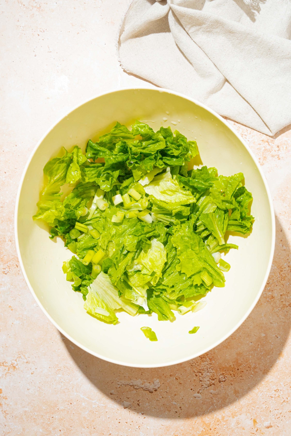 A large white bowl with chopped lettuce and sliced green onions. The bowl is on a tan counter with a white cloth napkin.