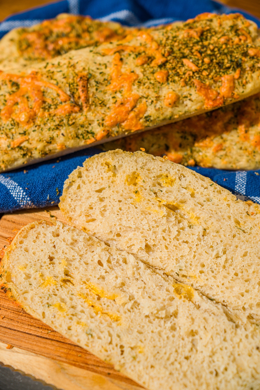 A cutting board with a baked Italian herb and cheese bread loaf sliced down the middle. The board is on a wooden counter with a basket of Italian bread loaves.