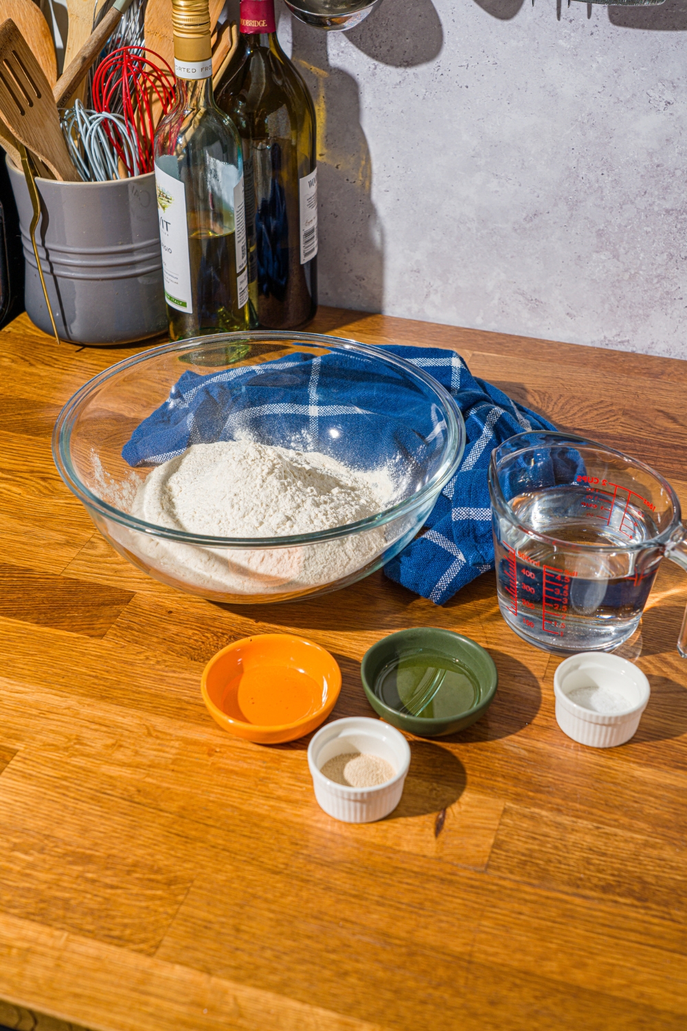 A wooden counter with ingredients to make Italian herb and cheese bread including water, flour, yeast, honey, and olive oil.