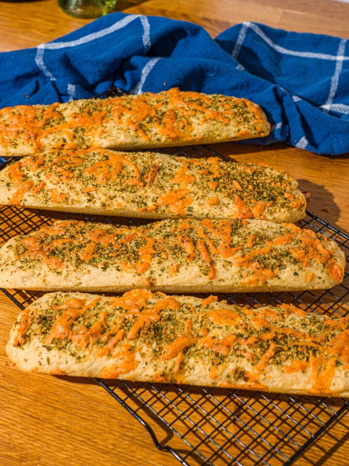 Four baked Italian herb and cheese bread loaves on a wire rack. The rack is on a wooden counter with a blue cloth napkin.