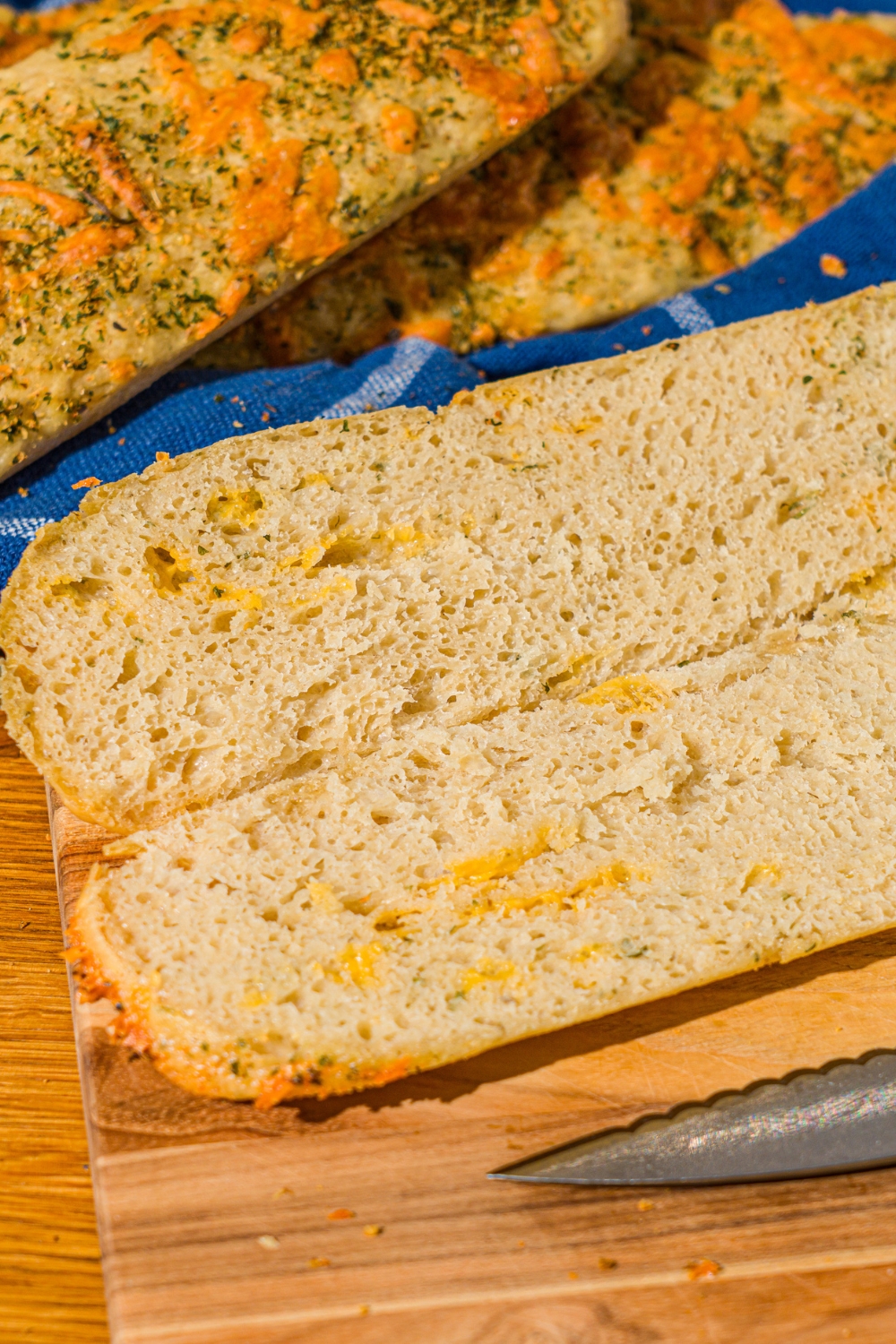 A cutting board with a baked Italian herb and cheese bread loaf sliced down the middle. The board is on a wooden counter with a basket of Italian bread loaves.