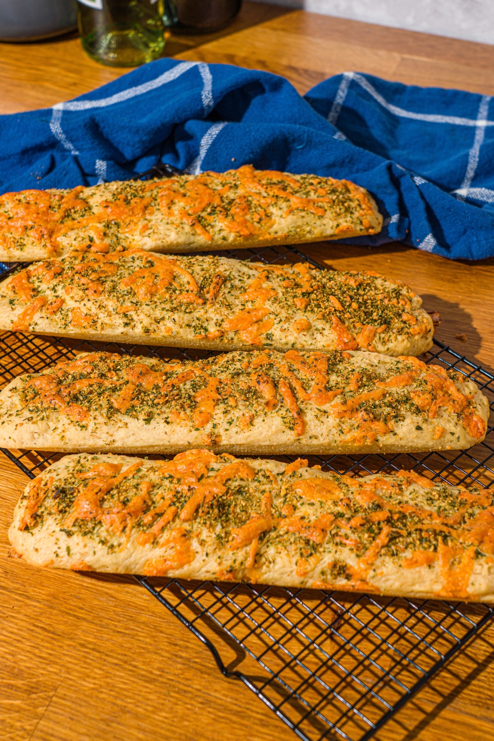 Four baked Italian herb and cheese bread loaves on a wire rack. The rack is on a wooden counter with a blue cloth napkin.