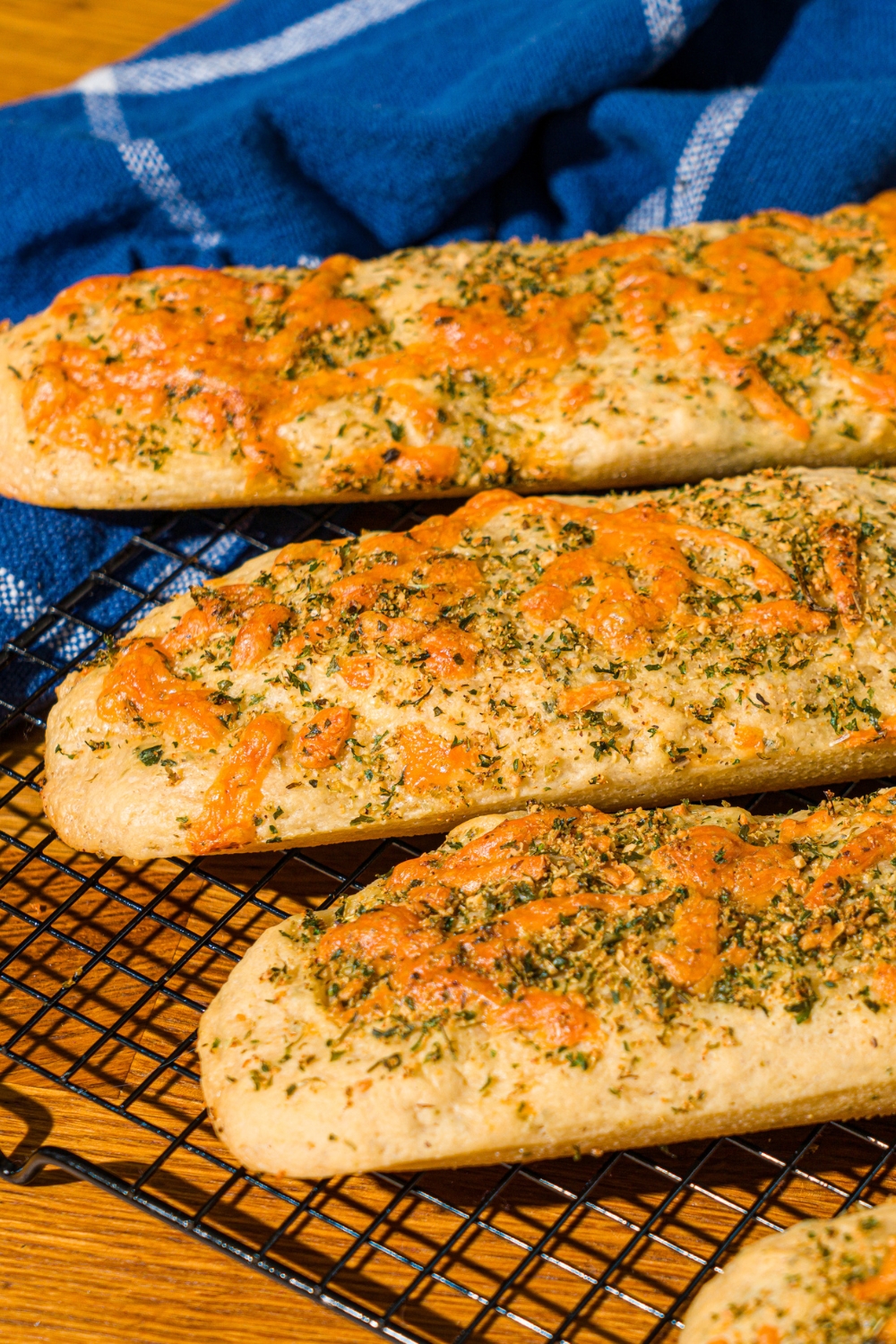 Four baked Italian herb and cheese bread loaves on a wire rack. The rack is on a wooden counter with a blue cloth napkin.