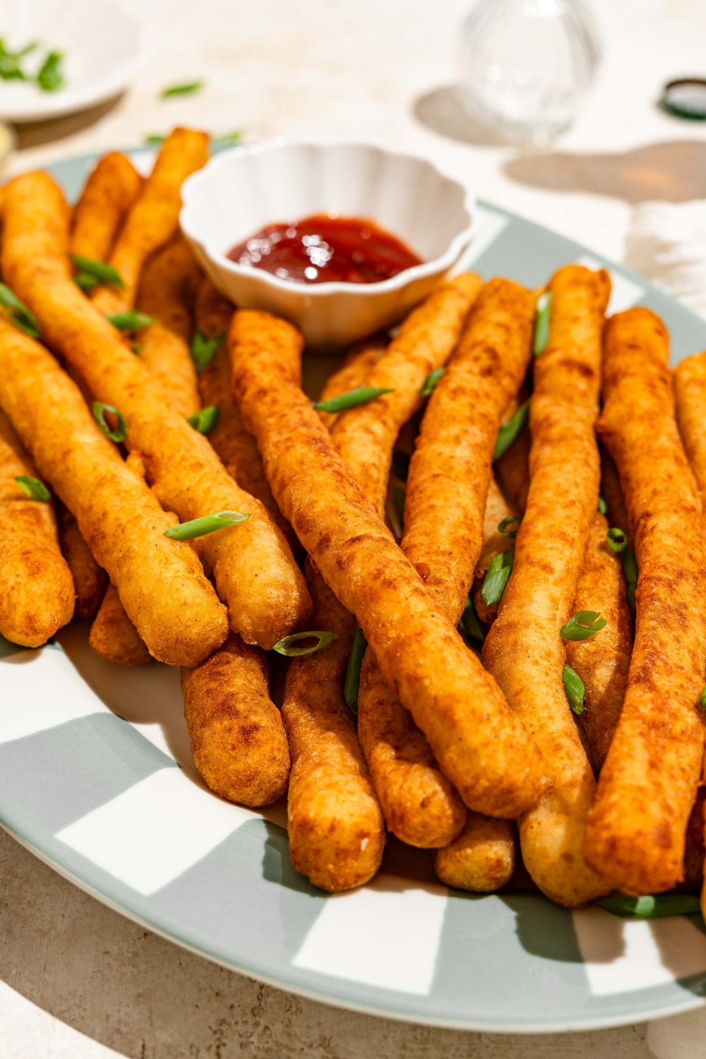 A striped plate with fried Japanese long fries garnished with sliced green onion and served with a small bowl of ketchup. The plate is on a tan counter.