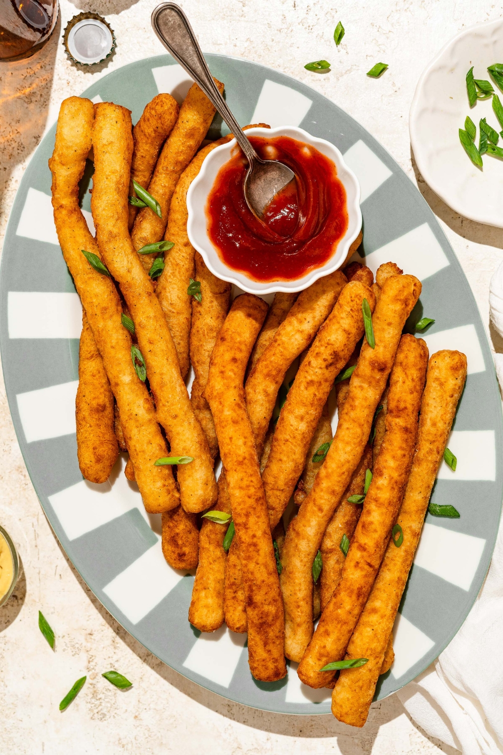 A striped plate with fried Japanese long fries garnished with sliced green onion and served with a small bowl of ketchup. The plate is on a tan counter.