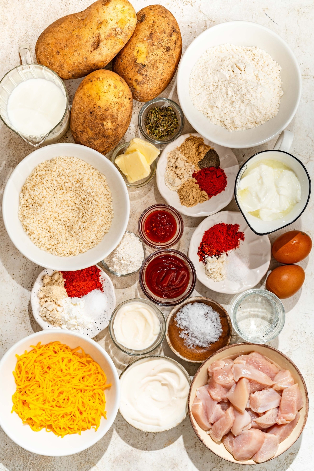 A tan counter with several bowls containing ingredients to make loaded fries including potatoes, chicken, shredded cheese, milk, flour, breadcrumbs, yogurt, ketchup, eggs, butter, mayo, herbs, and seasonings.