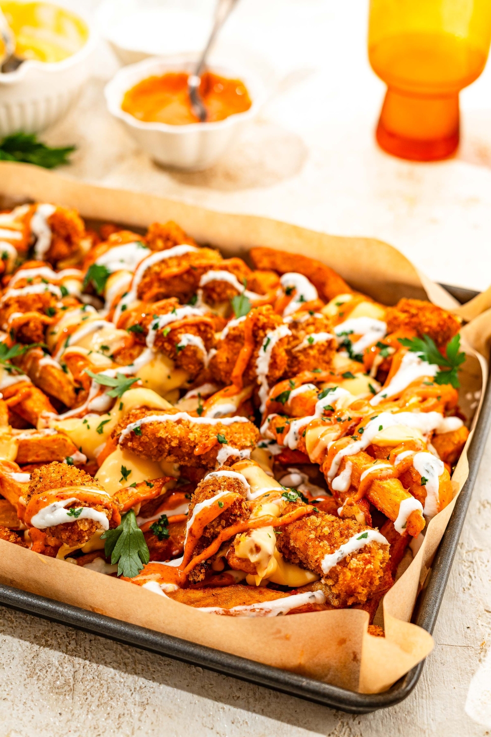 A baking sheet lined with parchment paper with loaded fries topped with crispy chicken, sriracha mayo sauce, yogurt sauce, cheese sauce, and fresh parsley. The sheet is on a tan counter with bowls of toppings.