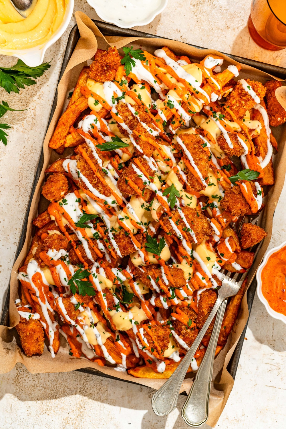 A baking sheet lined with parchment paper with loaded fries topped with crispy chicken, sriracha mayo sauce, yogurt sauce, cheese sauce, and fresh parsley. There are a pair of forks in the fries. The sheet is on a tan counter with bowls of toppings.