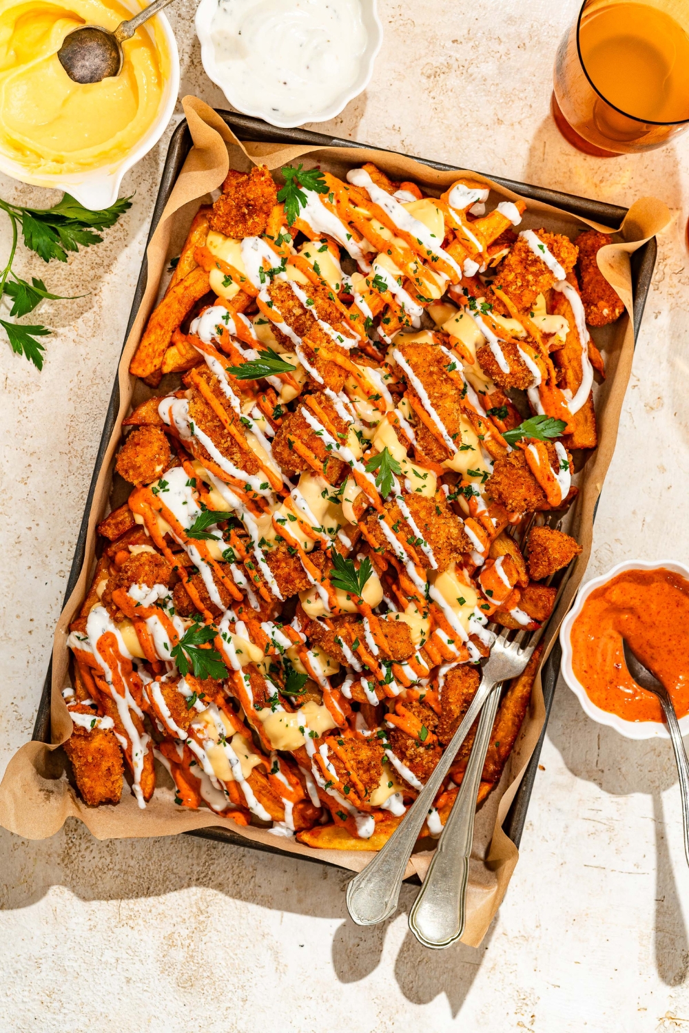 A baking sheet lined with parchment paper with loaded fries topped with crispy chicken, sriracha mayo sauce, yogurt sauce, cheese sauce, and fresh parsley. There are a pair of forks in the fries. The sheet is on a tan counter with bowls of toppings.