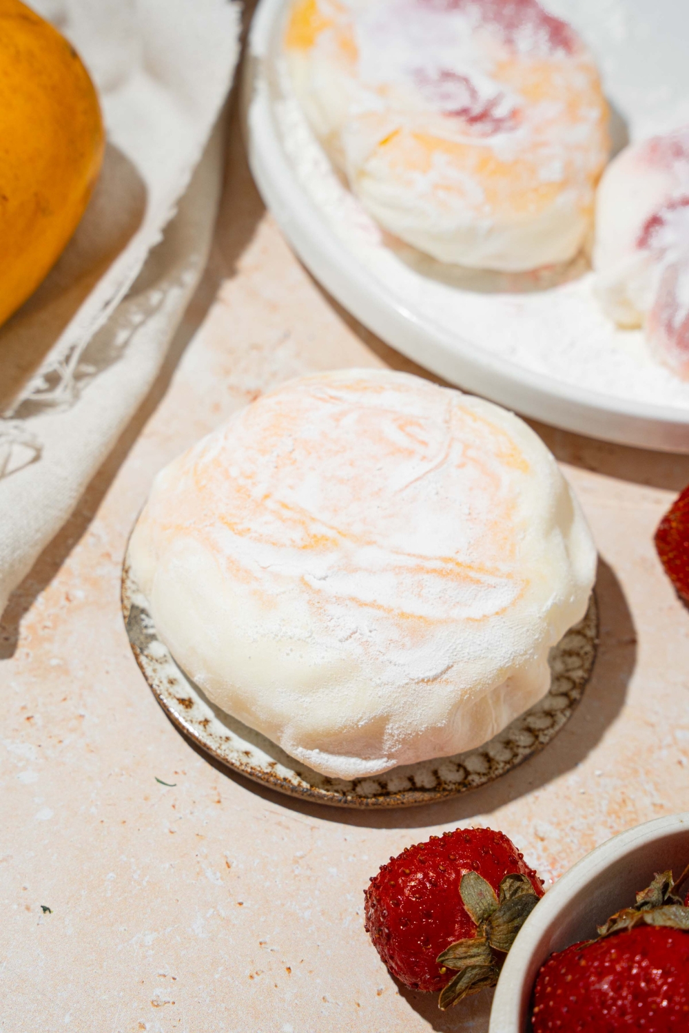 A plate with mango and strawberry rice paper mochi coated in rice flour. The plate is on a tan counter with an additional plate of mochis and a bowl of strawberries.