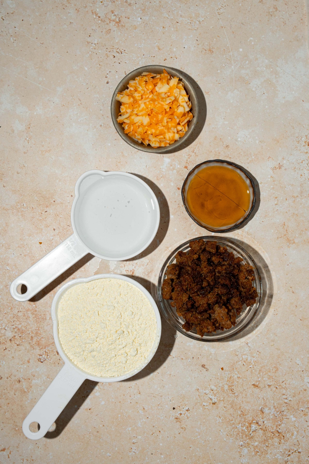 A tan counter with several bowls containing ingredients to make McGriddle muffins including pancake mix, breakfast sausage, shredded cheese, syrup, and water.