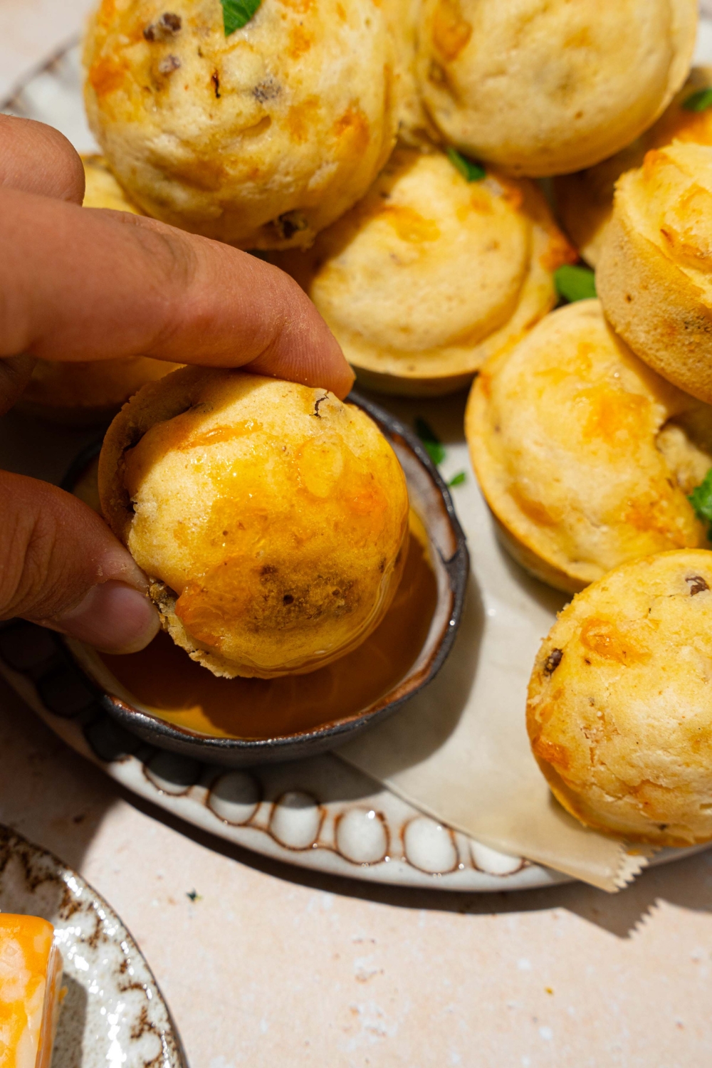 A hand dipping a McGriddle muffin into a small bowl of syrup. The bowl is on a plate lined with parchment paper and a pile of McGriddle muffins.