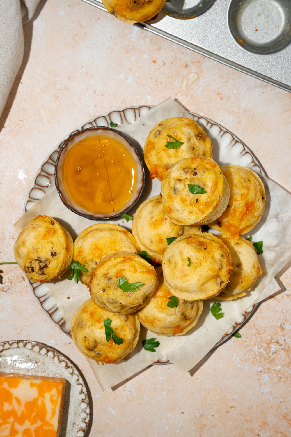 A plate lined with parchment paper with a pile of McGriddle muffins. The plate is garnished with fresh parsley and served with a small bowl of syrup. The plate is on a tan counter with a plate of cheese.