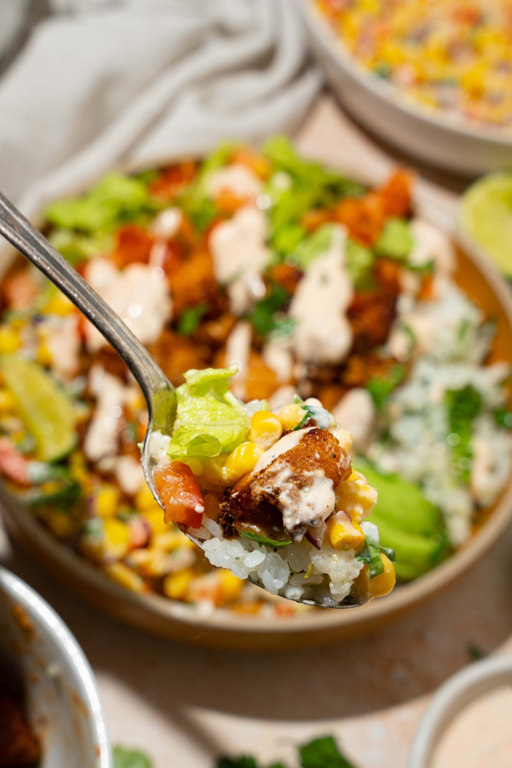 A close up of a fork with a bite of street corn chicken rice bowl. There is a rice bowl with cilantro rice, seasoned chicken, street corn, avocado, and a greek yogurt dressing blurred in the background.