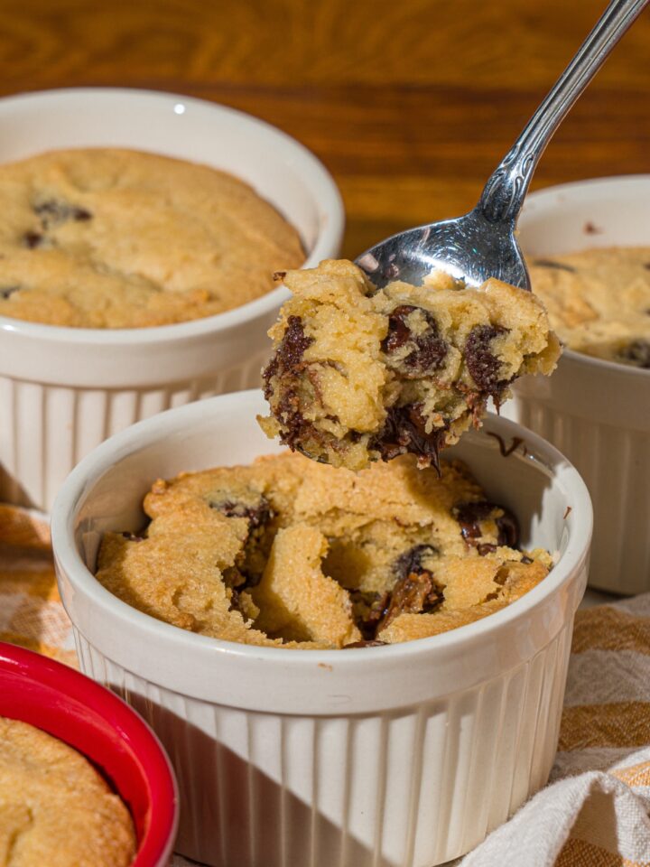 A ramekin with nutella cookie pie with a spoon taking a bite from the pie. The ramekin is on a wooden counter with additional pies.