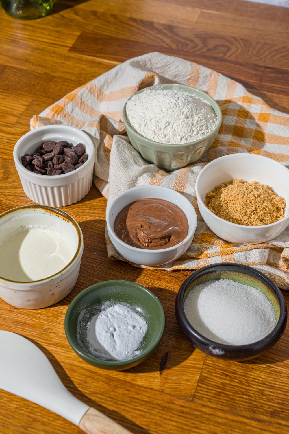 A wooden counter with several bowls of ingredients to make nutella cookie pie including nutella, flour, butter, sugar, brown sugar, chocolate chips, nad more.