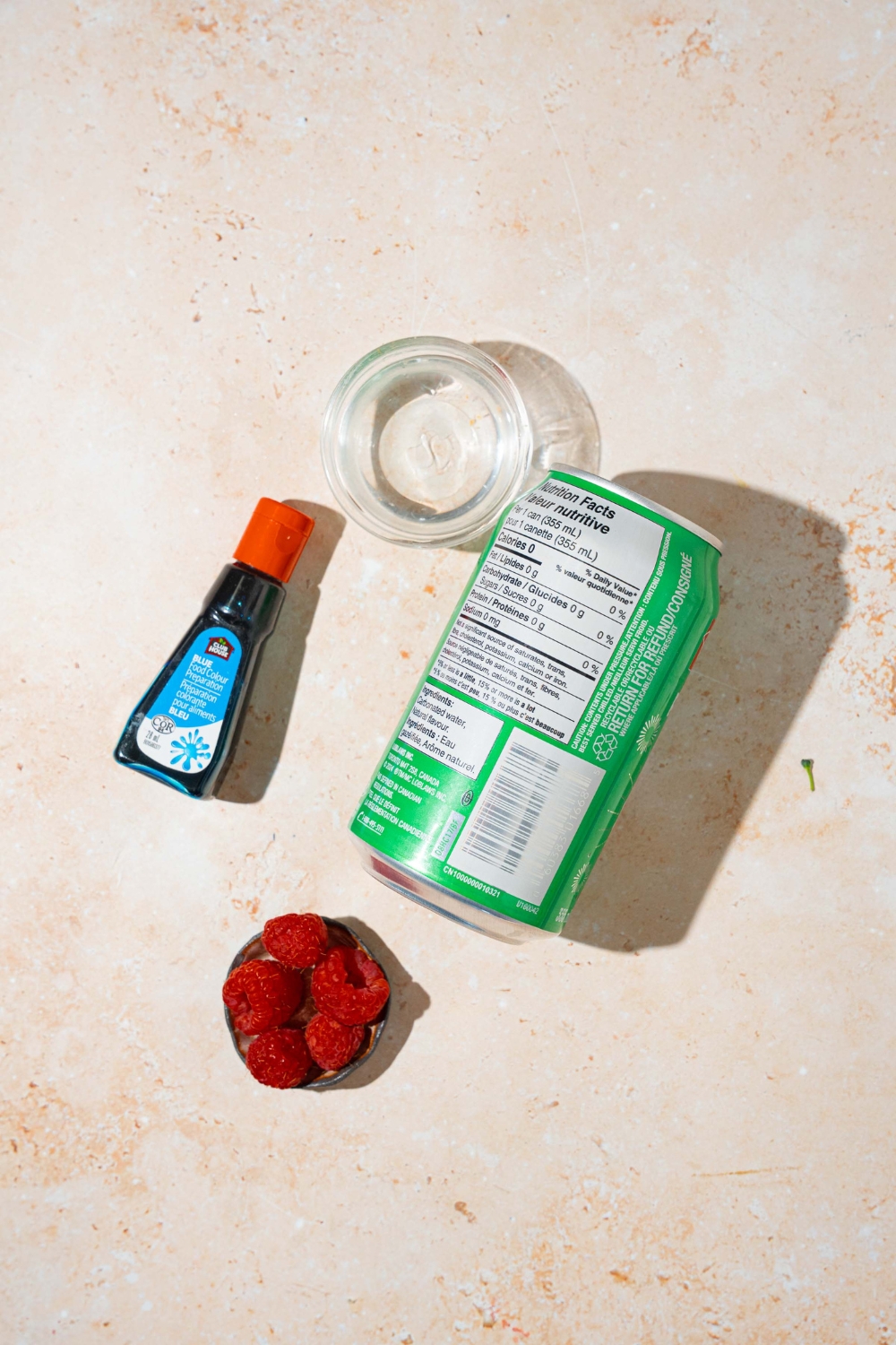 A tan counter with ingredients to make ocean water drink including a can of Sprite, coconut syrup, Blue food coloring, and raspberries.