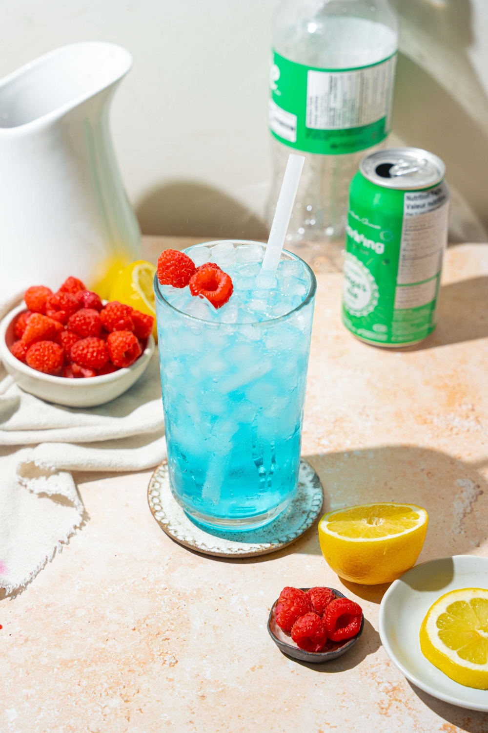 A tall glass of ocean water drink served with pebble ice, a straw, and garnished with fresh raspberries. The glass is on a coaster on a tan counter with a bottle and can of soda, bowls of raspberries, and sliced lemon.