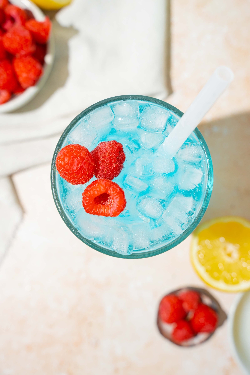 A tall glass of ocean water drink served with pebble ice, a straw, and garnished with fresh raspberries. The glass is on a coaster on a tan counter with a bottle and can of soda, bowls of raspberries, and sliced lemon.