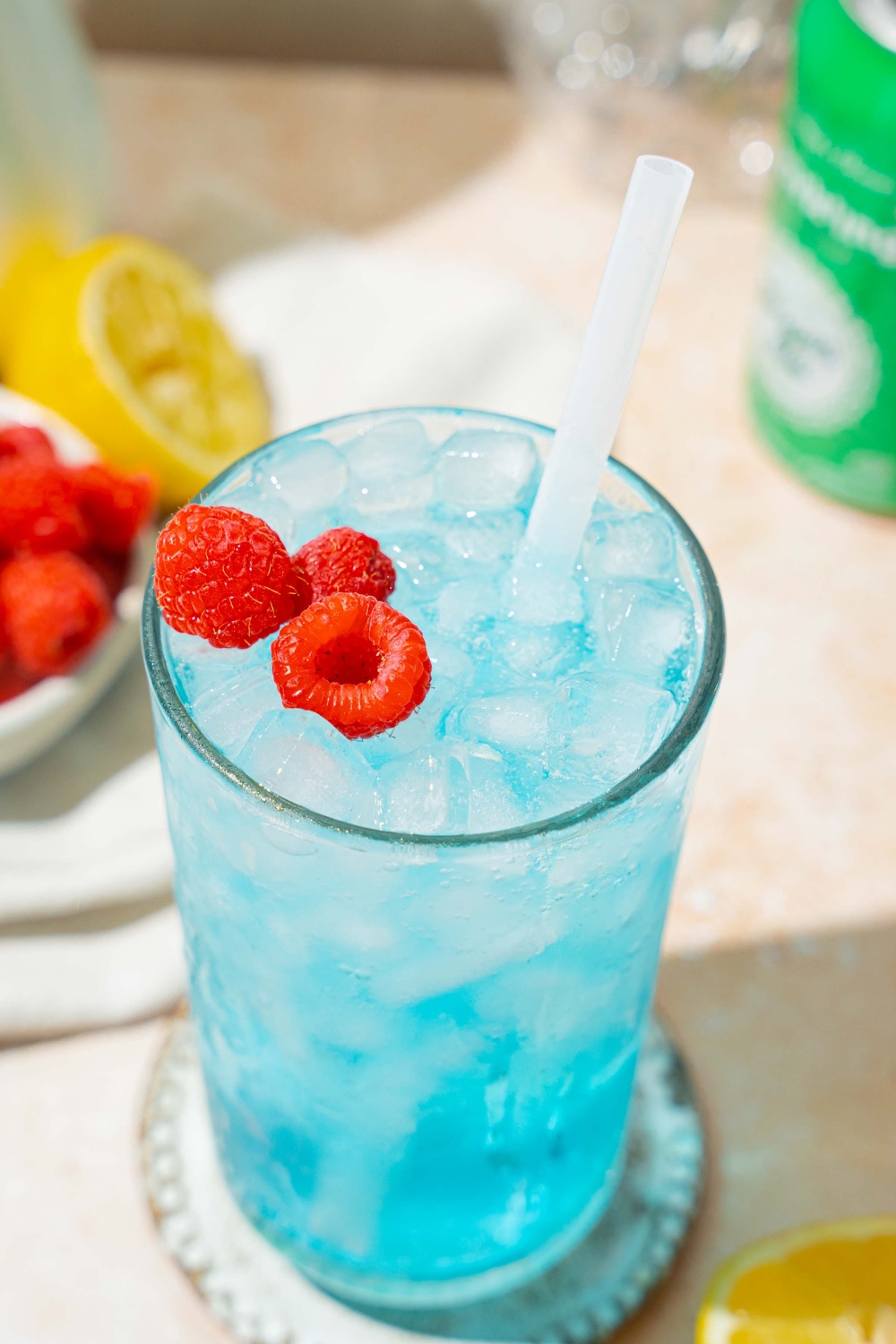 A tall glass of ocean water drink served with pebble ice, a straw, and garnished with fresh raspberries. The glass is on a coaster on a tan counter with a bottle and can of soda, bowls of raspberries, and sliced lemon.