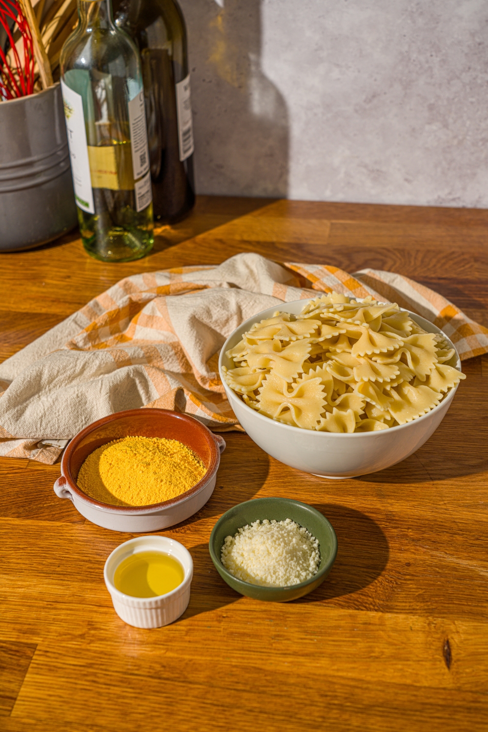A wooden counter with ingredients to make pasta chips including a bowl of bowtie pasta, oil, parmesan cheese, mac and cheese powder, and a yellow striped napkin.