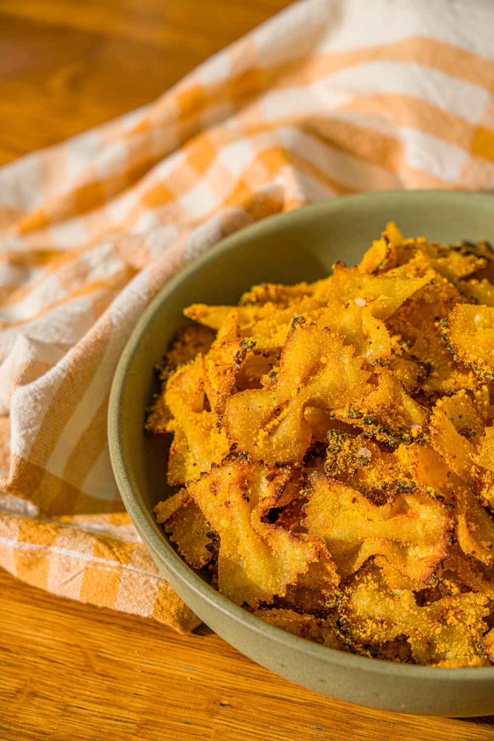 A green bowl with air fried bowtie pasta chips tossed in parmesan and mac and cheese powder. The bowl is on a wooden counter with a yellow striped napkin.