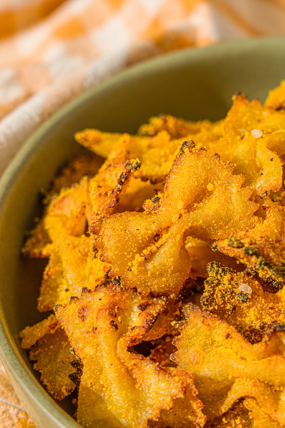 A green bowl with air fried bowtie pasta chips tossed in parmesan and mac and cheese powder. The bowl is on a wooden counter with a yellow striped napkin.