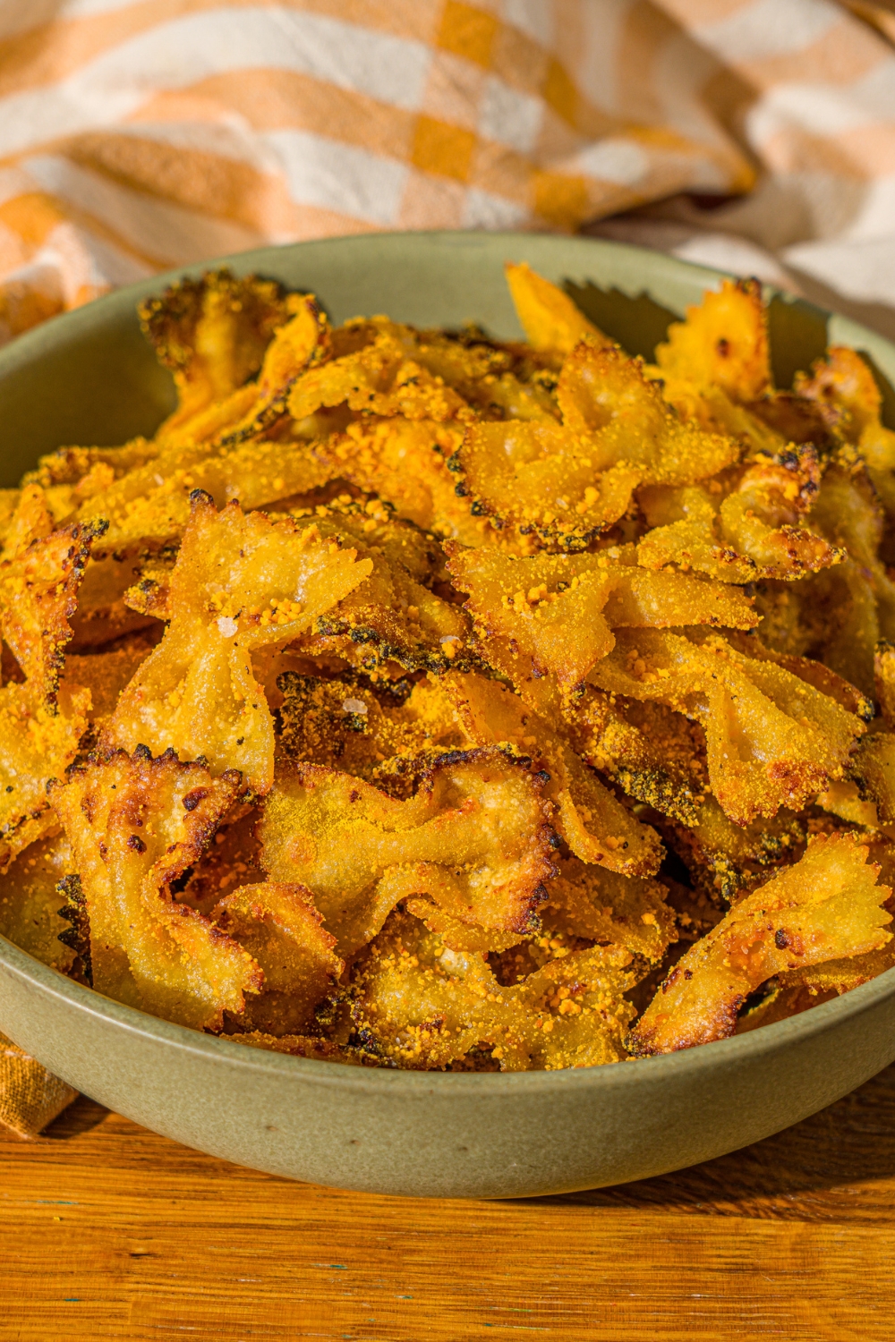 A green bowl with air fried bowtie pasta chips tossed in parmesan and mac and cheese powder. The bowl is on a wooden counter with a yellow striped napkin.