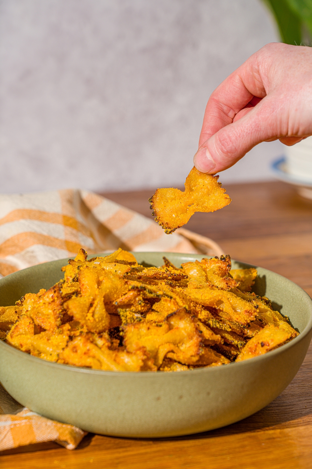 A bowl of bowtie pasta chips seasoned with cheese. There is a hand taking a pasta chip from the bowl. The bowl is on a wooden counter with a yellow striped napkin.