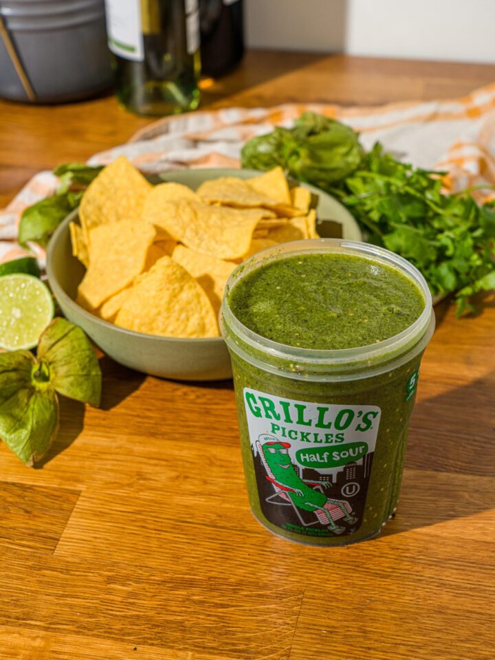 A wooden counter with pickle salsa stored in a Grillo's pickles and a bowl of tortilla chips.