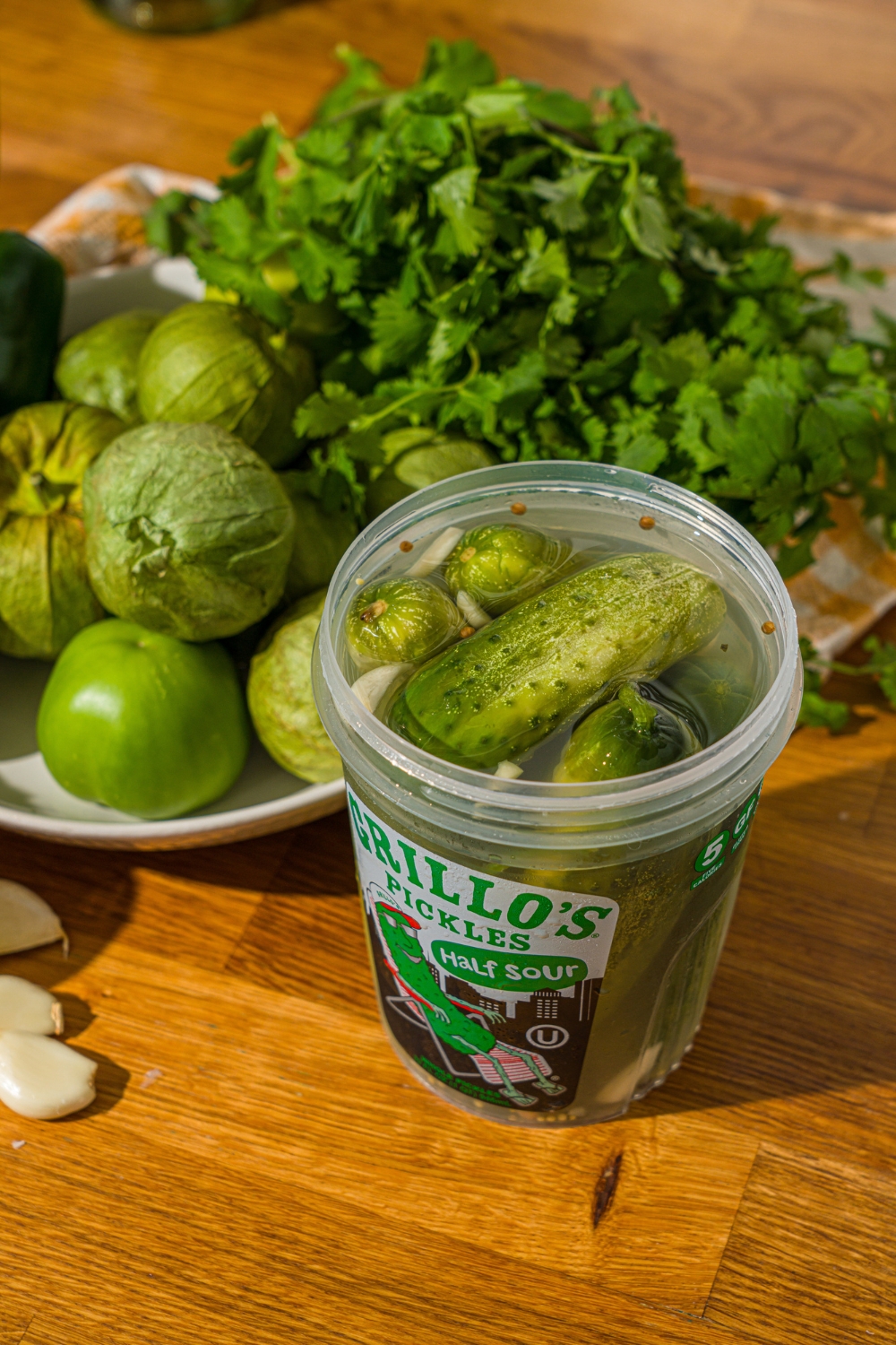 A wooden counter with a container of Grillo's pickles and a white bowl with poblano peppers, lime, tomatillos, and cilantro.