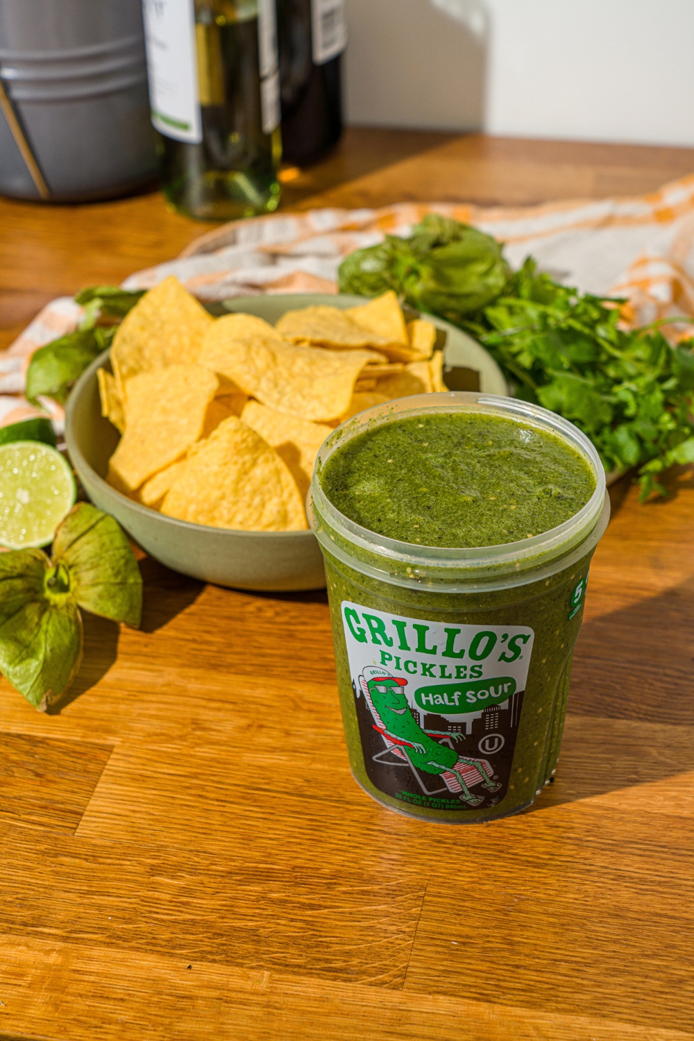 A wooden counter with pickle salsa stored in a Grillo's pickles and a bowl of tortilla chips.