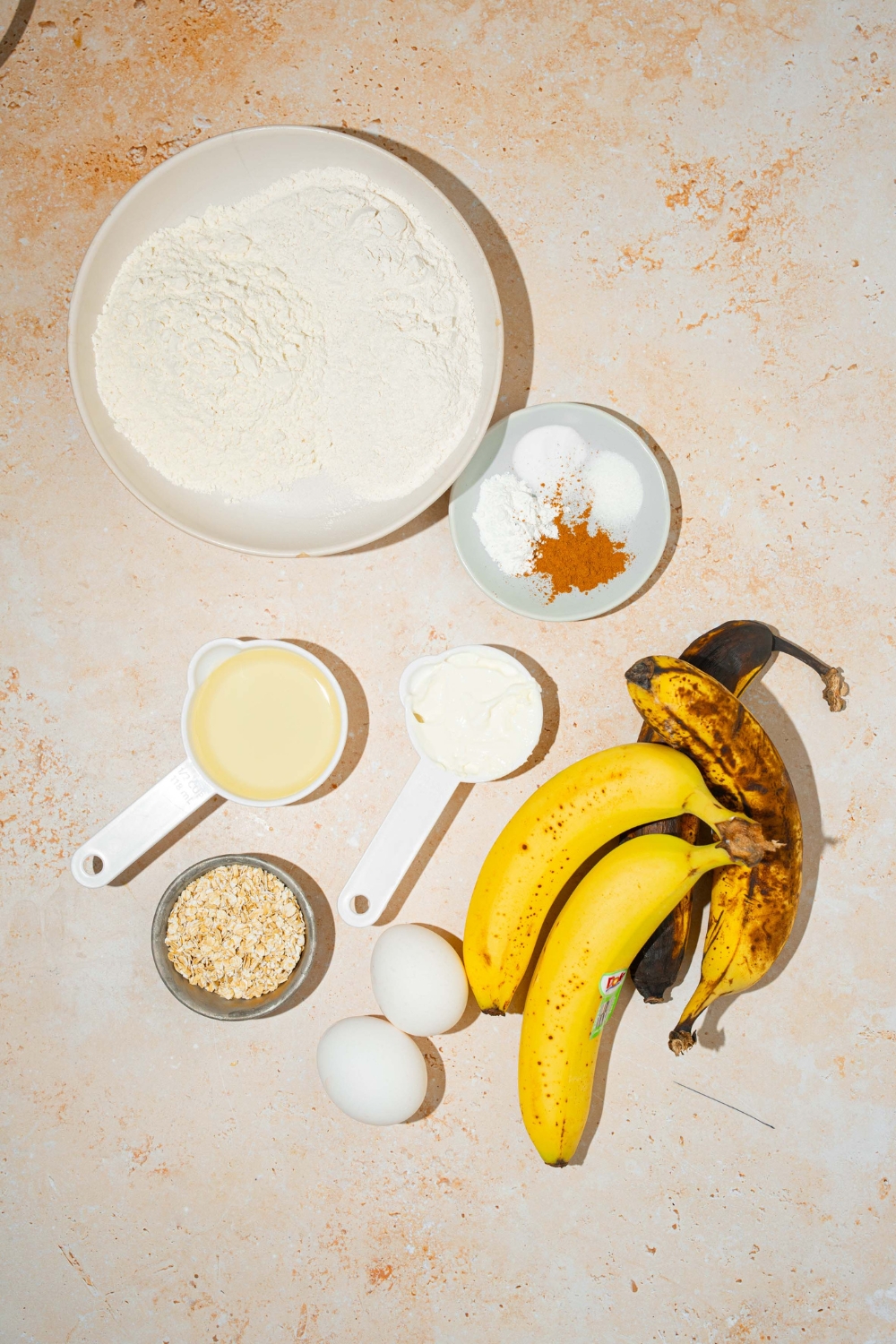 A bowl of flour, a small plate with cinnamon, baking powder, baking soda, and salt on it, a cup of oil, a cup of sour cream, a bowl of oats, four bananas and two eggs all on a white counter.