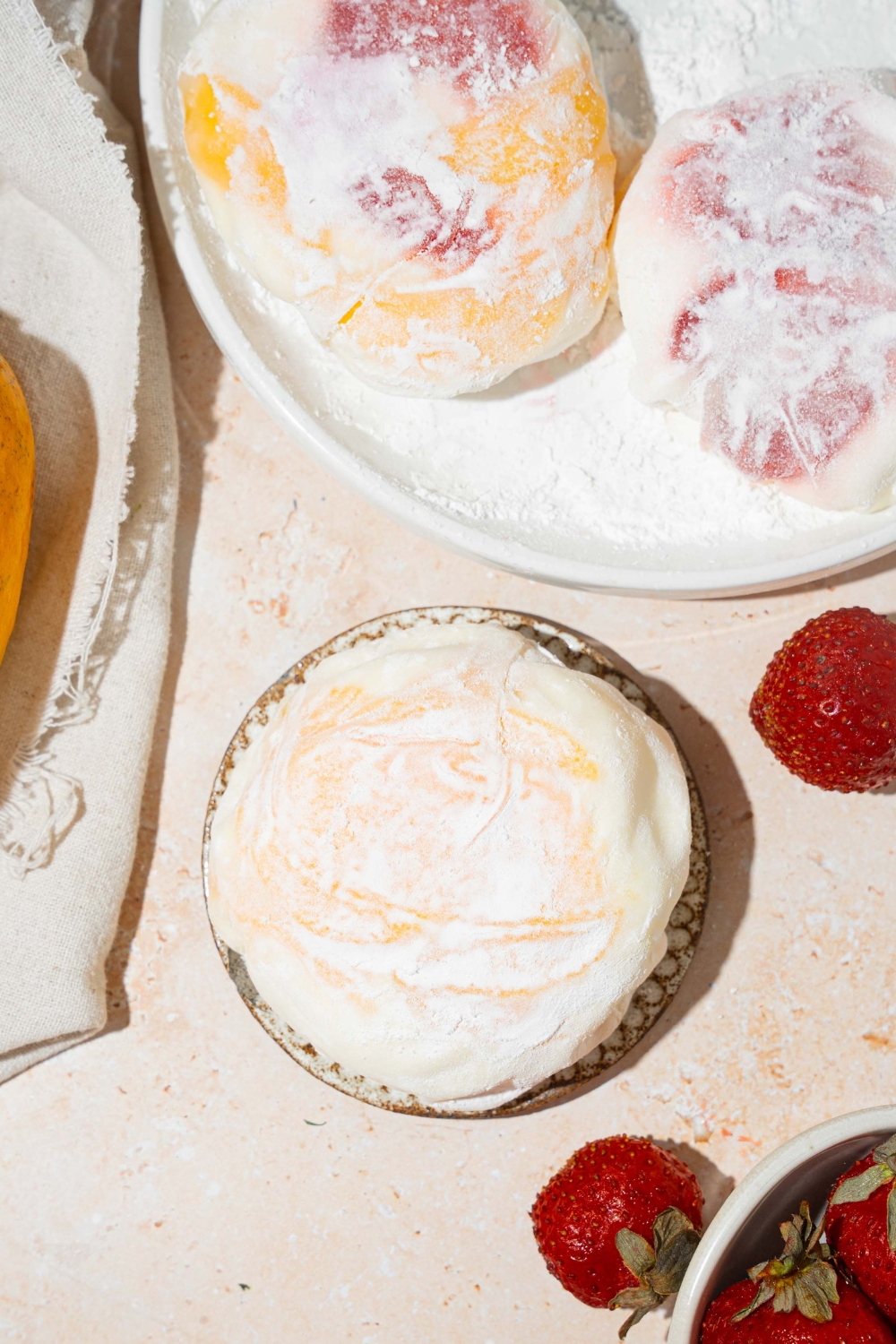 A plate with mango and strawberry rice paper mochi coated in rice flour. The plate is on a tan counter with an additional plate of mochis and a bowl of strawberries.